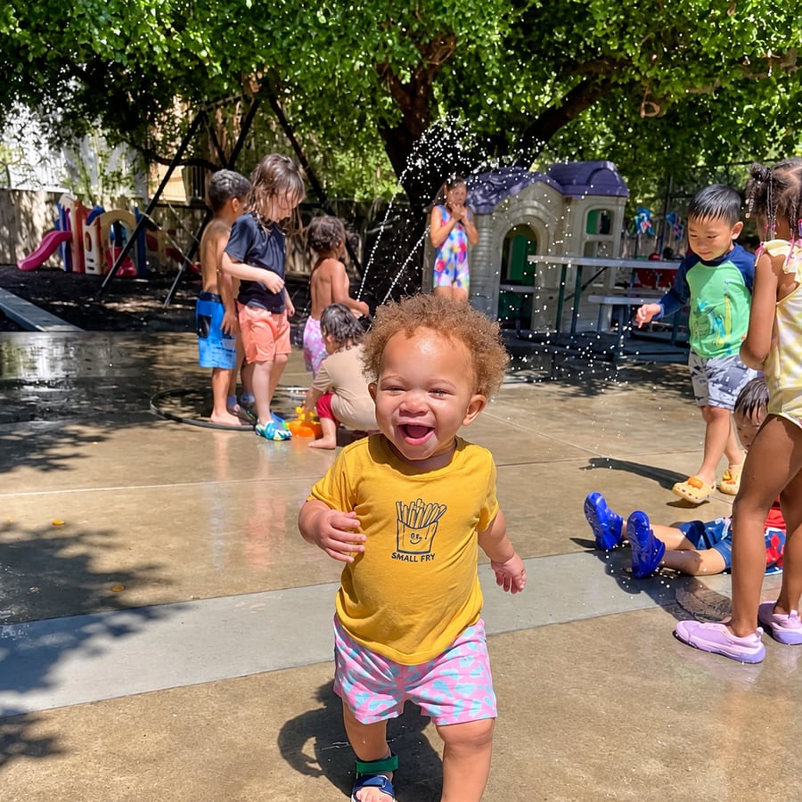 A laughing toddler running through sprinklers on the play yard