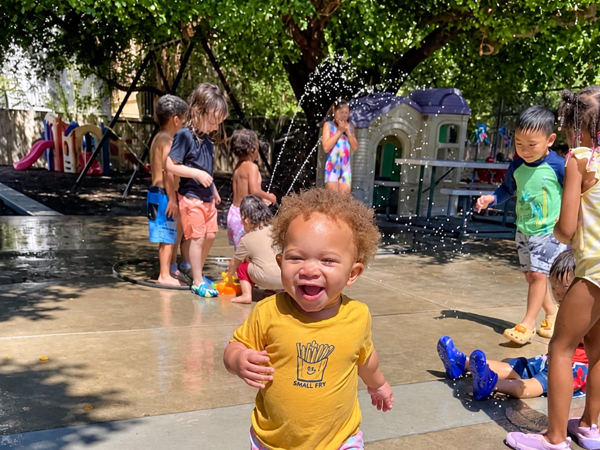 A laughing toddler running through sprinklers on the play yard