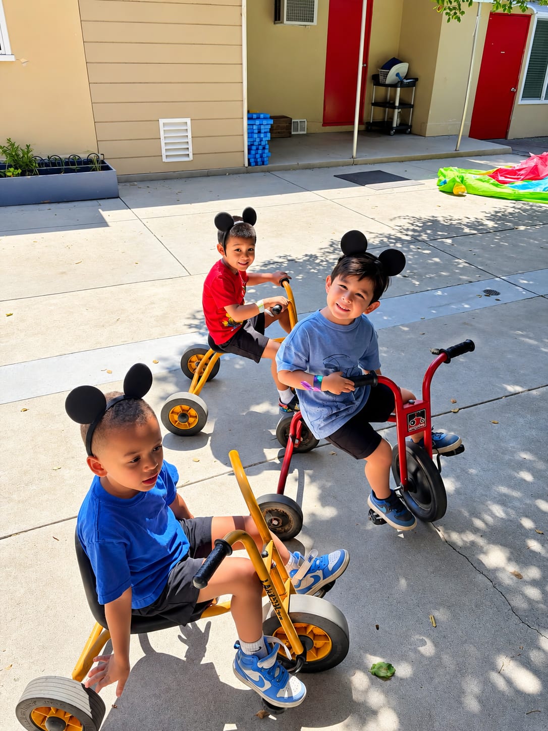 Three boys in Mickey Mouse ears riding tricycles on the campus walkway