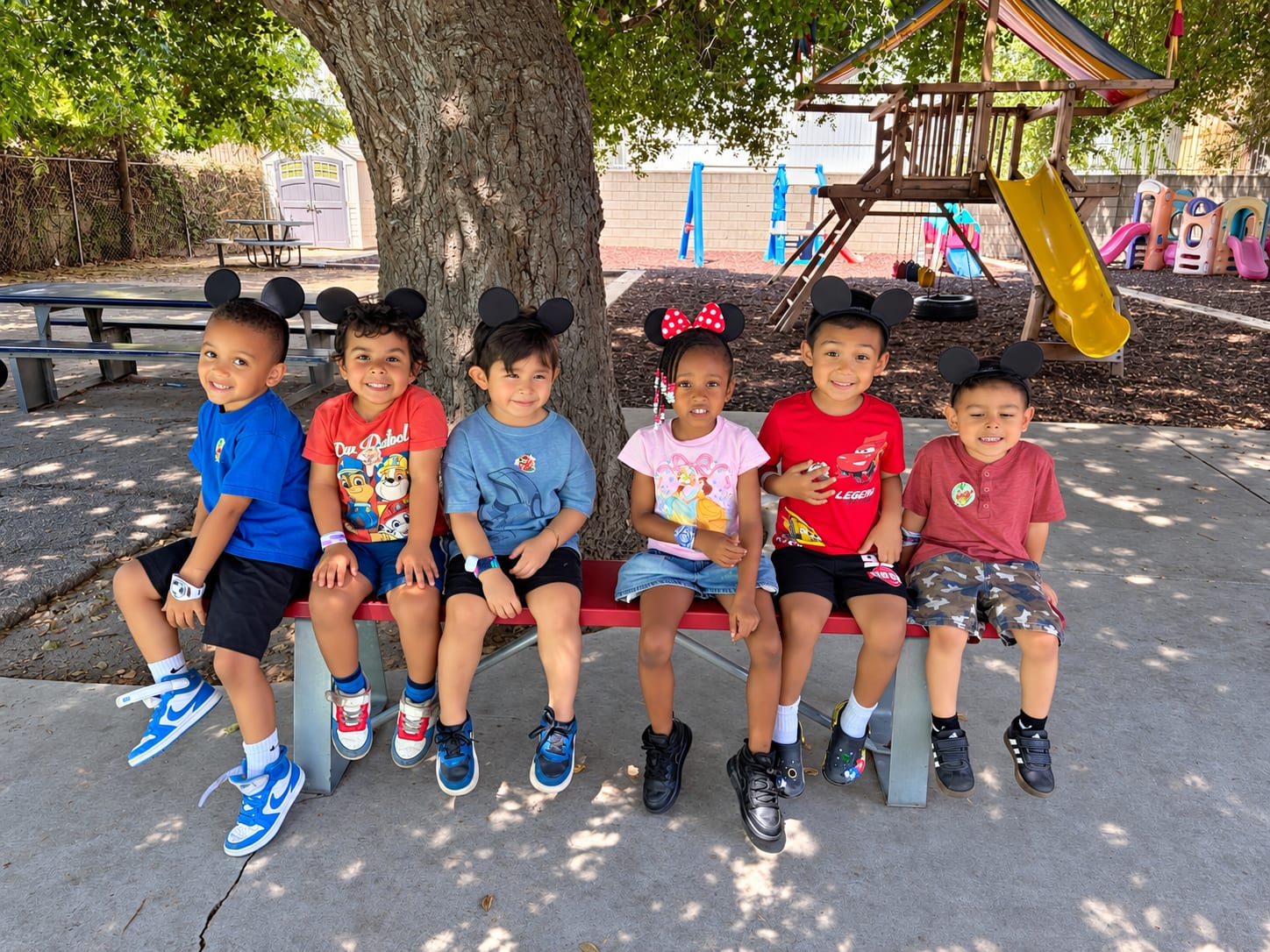 A group of six preschoolers in Mickey ears smiling on a bench in the play yard